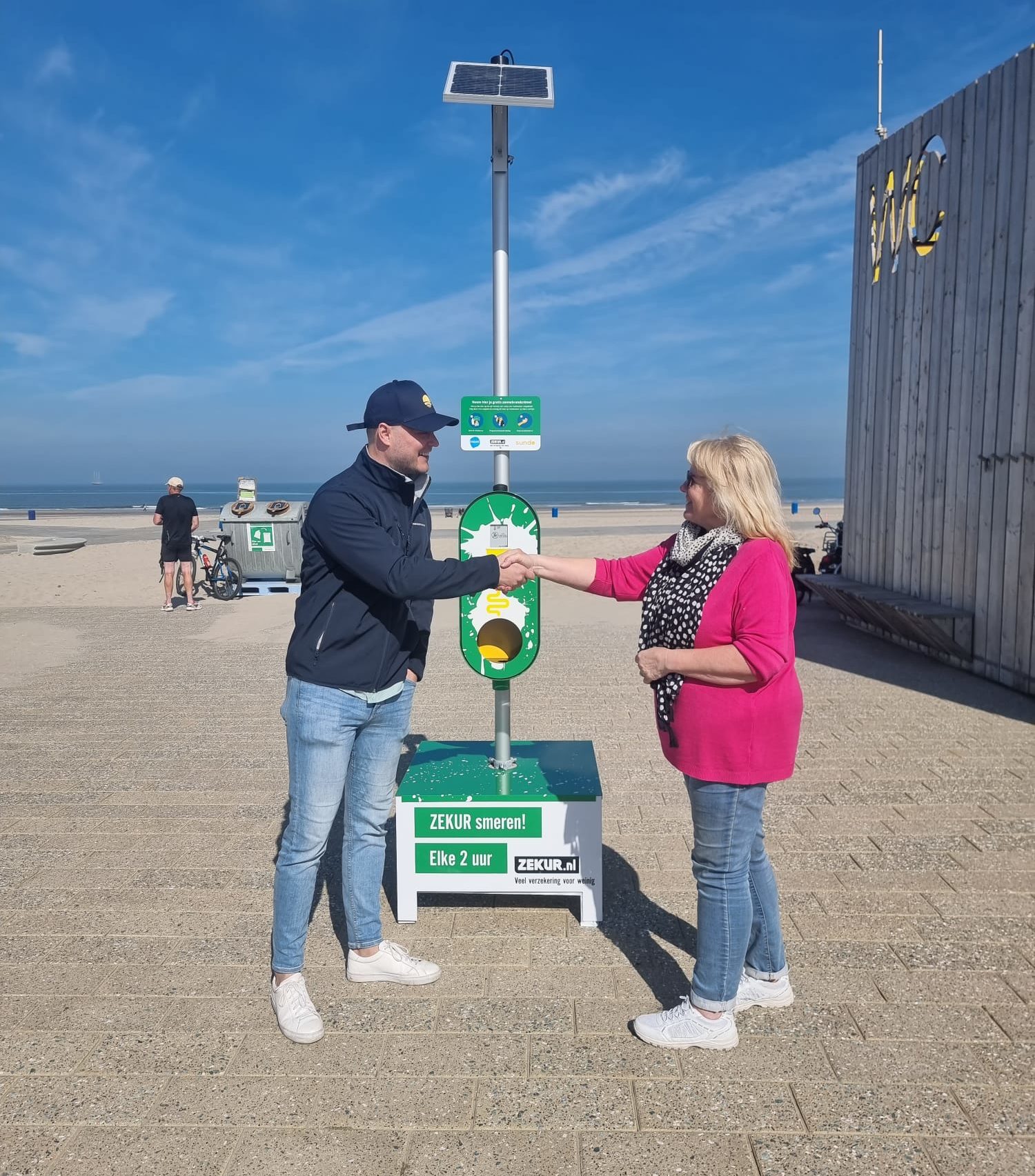 Ybe Heemskerk (Sundo) en Josje van den Hoonaard (strandcoördinator) schudden elkaar de hand voor de zonnebrandpaal van Hoek van Holland