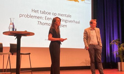 Twee personen op een podium voor een presentatiedia met de tekst 'Het taboe op mentale problemen. Een ervaringsverhaal'
