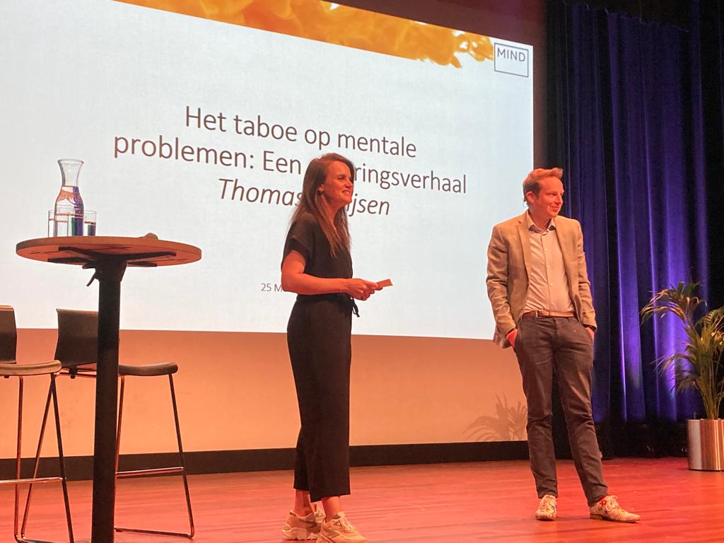 Twee personen op een podium voor een presentatiedia met de tekst 'Het taboe op mentale problemen. Een ervaringsverhaal'