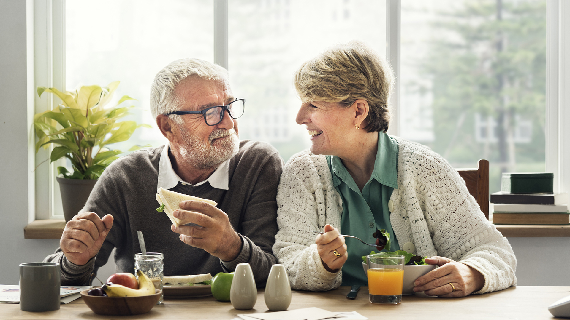 Oudere man en vrouw aan de keukentafel met ontbijt, kijken elkaar blij aan