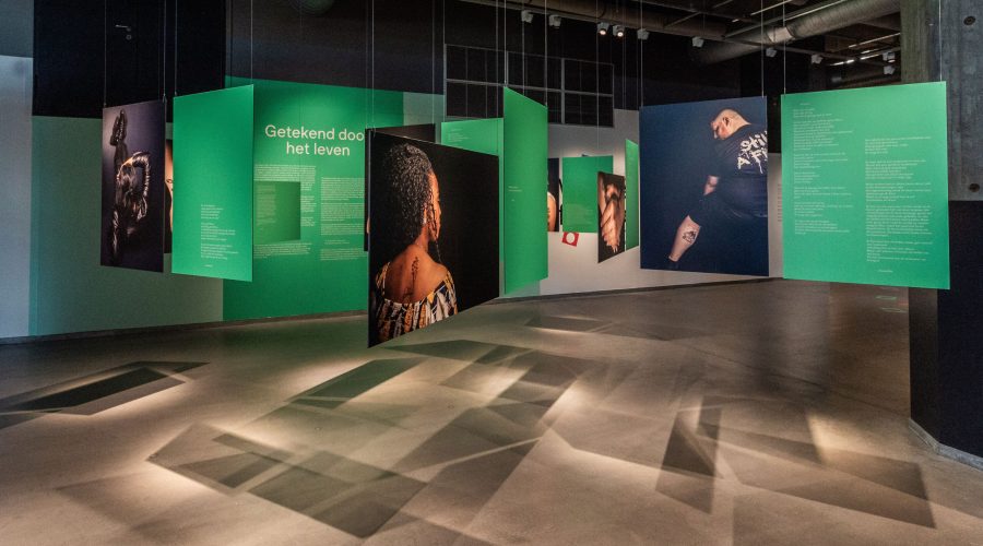 Foto van de expositie waarbij je verschillende foto's ziet hangen in een ruimte, met daarbij ook groene vlakken met een toelichting erop.