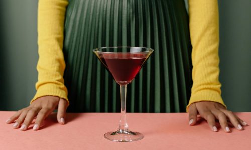 Cocktail glas met alcohol op een tafel. Daarachter zie je de armen en rok van een vrouw.