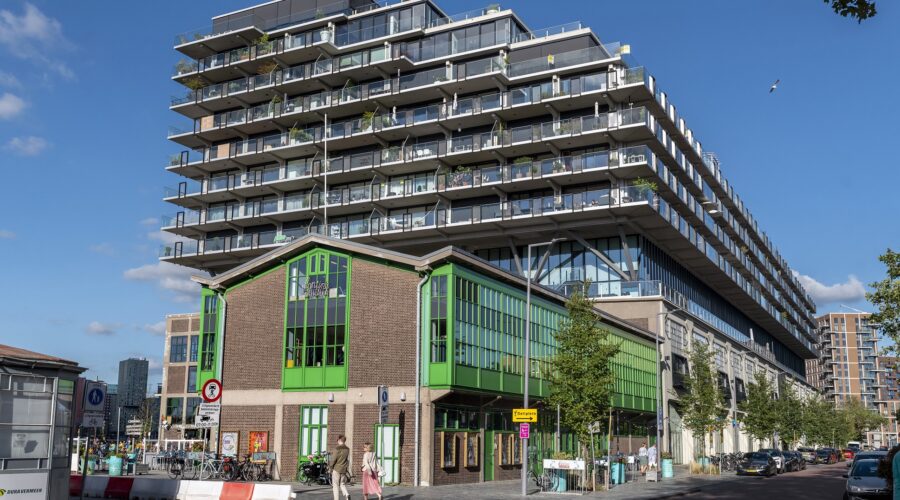 Foto van kantine Walhalla in Rotterdam. Het is een industrieel gebouw van bakstenen en groene kozijnen. Je ziet op de achtergrond een groot flatgebouw.